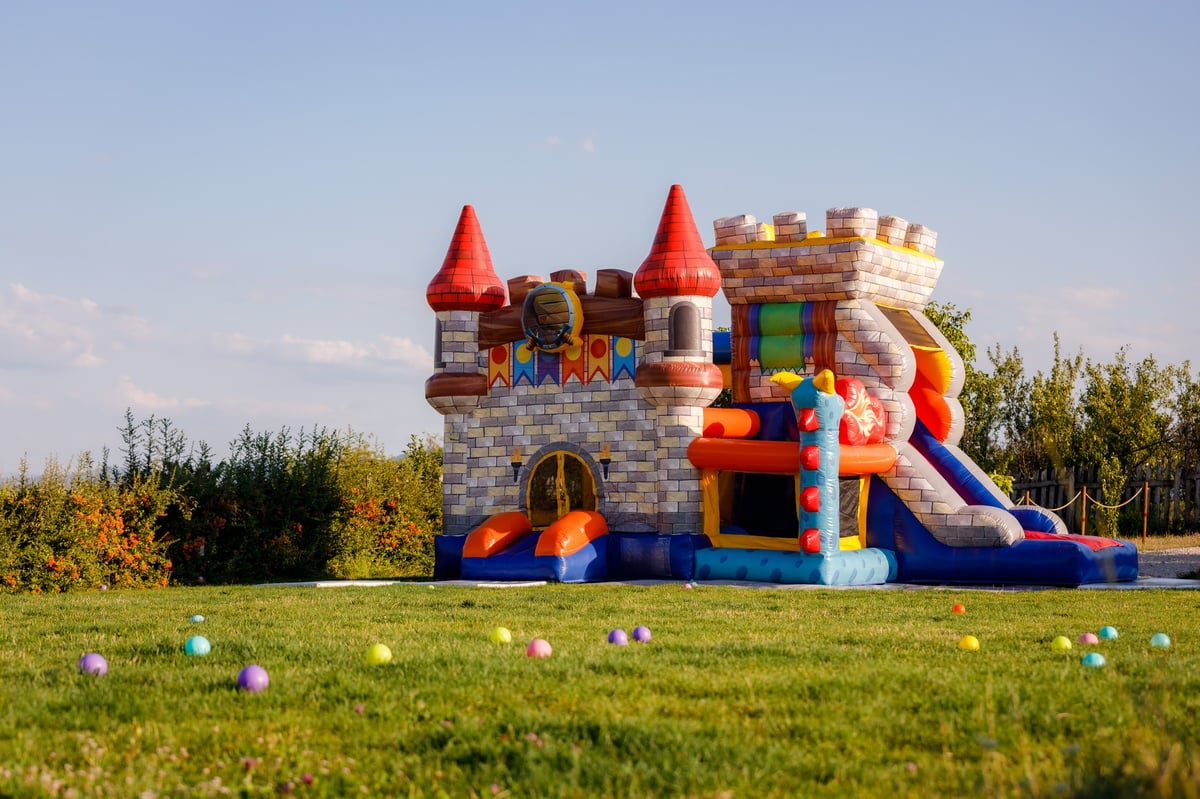 A vibrant inflatable castle bounce house sits in a lush green field under a clear blue sky, surrounded by colorful balls scattered on the grass, created as an independent concept for a Party & Event Rentals website