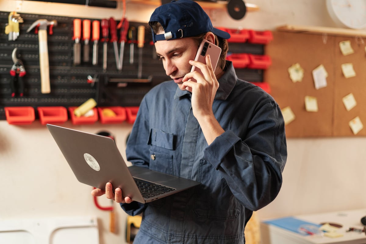 Busy male manual worker using laptop and talking on phone in woodworking shop with tools on wall, discussing a project at his workplace