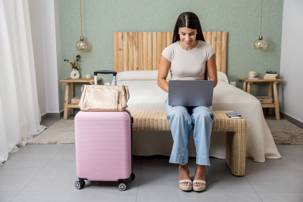 Young woman working on laptop beside suitcase in hotel room, created as an independent concept for an online booking website